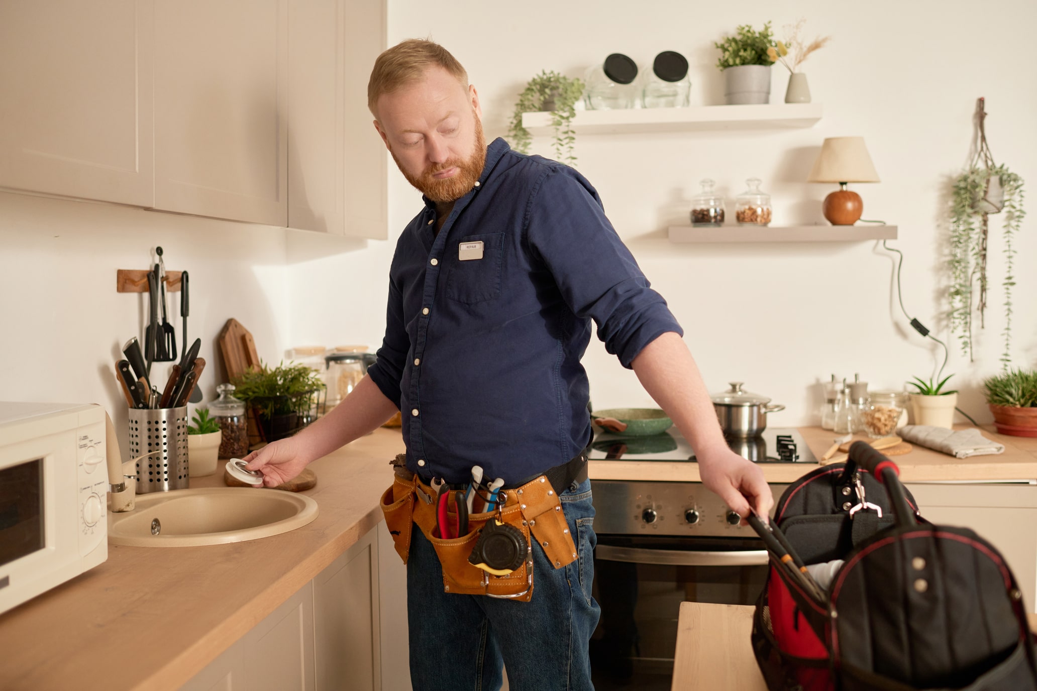 Man in a blue shirt and tool belt inspecting a kitchen sink.