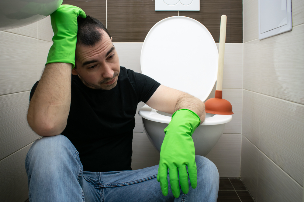 A man wearing green gloves sits on the floor next to a toilet, looking frustrated. A plunger is placed beside the toilet.